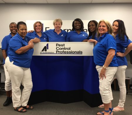 A1 Lynn Office staff photo, eight women pictured in company attire smiling surrounding A1 Pest Control Professionals sign.